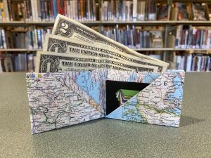 A wallet made of a folded atlas page holding a St Helens Public Library card, two $1 bills, and a $2 bill.